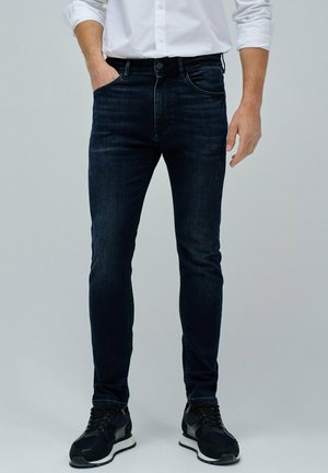 Jeans de mezclilla azul oscuro con un corte ajustado, cinco bolsillos y detalles de desvanecimiento sutil; combinados con zapatillas deportivas negras.