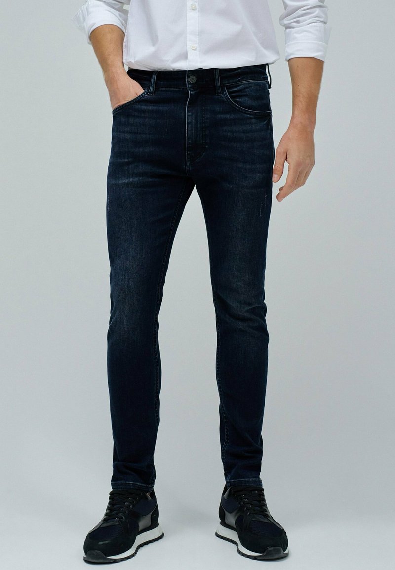 Jeans de mezclilla azul oscuro con un corte ajustado, cinco bolsillos y detalles de desvanecimiento sutil; combinados con zapatillas deportivas negras.