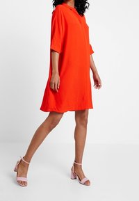 Vestito rosso maniche corte con una vestibilità rilassata, abbottonatura frontale e una texture liscia. Abbinato a sandali rosa con tacco a blocco.