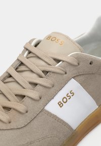 Beige semsket sko med hvite lærdetaljer, med gulllogo, rund tå, flate snørebånd og såle av gummigummi. Teksturerte overflater.