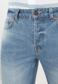 Vaqueros de mezclilla azul claro con un corte clásico, diseño de cinco bolsillos y cremallera. Presentan un desvanecimiento sutil y detalles metálicos en bronce.