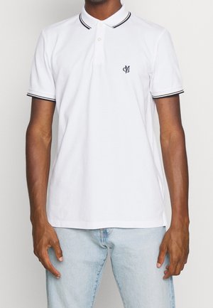 Man draagt een witte poloshirt met zwarte rand aan kraag en mouwen, gecombineerd met lichtblauwe spijkerbroek, staand tegen een effen achtergrond.