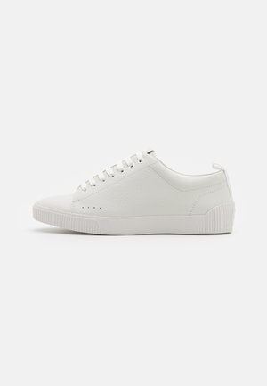 White leather sneaker with a textured finish, round toe, thin white laces, and a ribbed rubber sole. Features minimal perforations for breathability.