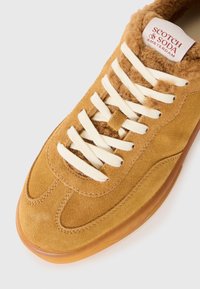 Brune suedesneakers med kremfargede snører og en myk, lys brun krage. Har en avrundet tå og en gjennomsiktig gummisåle. Logo på tungen.