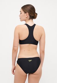 Svart badedrakt med racerback-top og lavt liv. Glatt tekstur med en liten gul logo på baksiden av underdelen.
