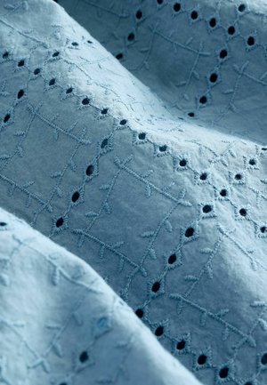 Lyseblåt stof med indviklet broderi, der viser mønstre af kors og små udskårede cirkler. Blød tekstur med et delikat design.