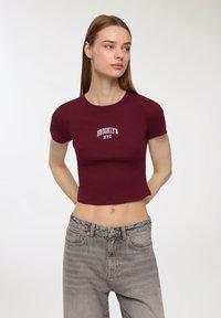 T-shirt corta bordeaux con maniche corte, caratterizzata dal testo bianco "BROOKLYN NYC". Abbinata a jeans grigi a vita alta con una texture sbiadita.