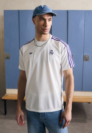 Biela športová dres s fialovými pruhmi, logo Adidas a emblém Real Madrid. V kombinácii s modrými džínsami a svetlomodrou čiapkou.