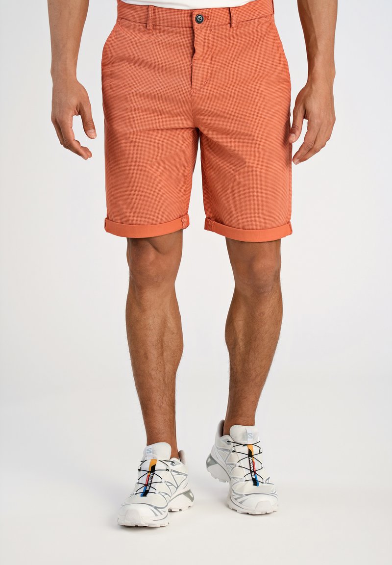 Pantaloni corti a quadretti arancioni con risvolti, dotati di chiusura con bottone e zip. Indossati con scarpe da ginnastica bianche e grigie con lacci colorati.