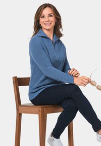 Blauer, strukturierter Pullover mit halbem Reißverschluss. Kombiniert mit dunklen Jeans und weißen Sneakers. Modell sitzt auf einem Holzstuhl und hält Sonnenbrillen.