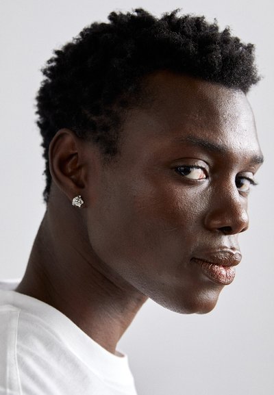 Modelo con cabello corto y texturizado negro, llevando un pendiente de plata. Piel suave con un resplandor natural, mirando ligeramente hacia la izquierda sobre un fondo claro.