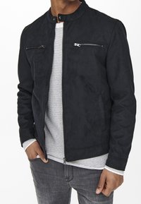 Giacca nera in suede con colletto alto, due tasche sul petto con zip e una trama liscia, abbinata a una camicia bianca a trama sottostante.