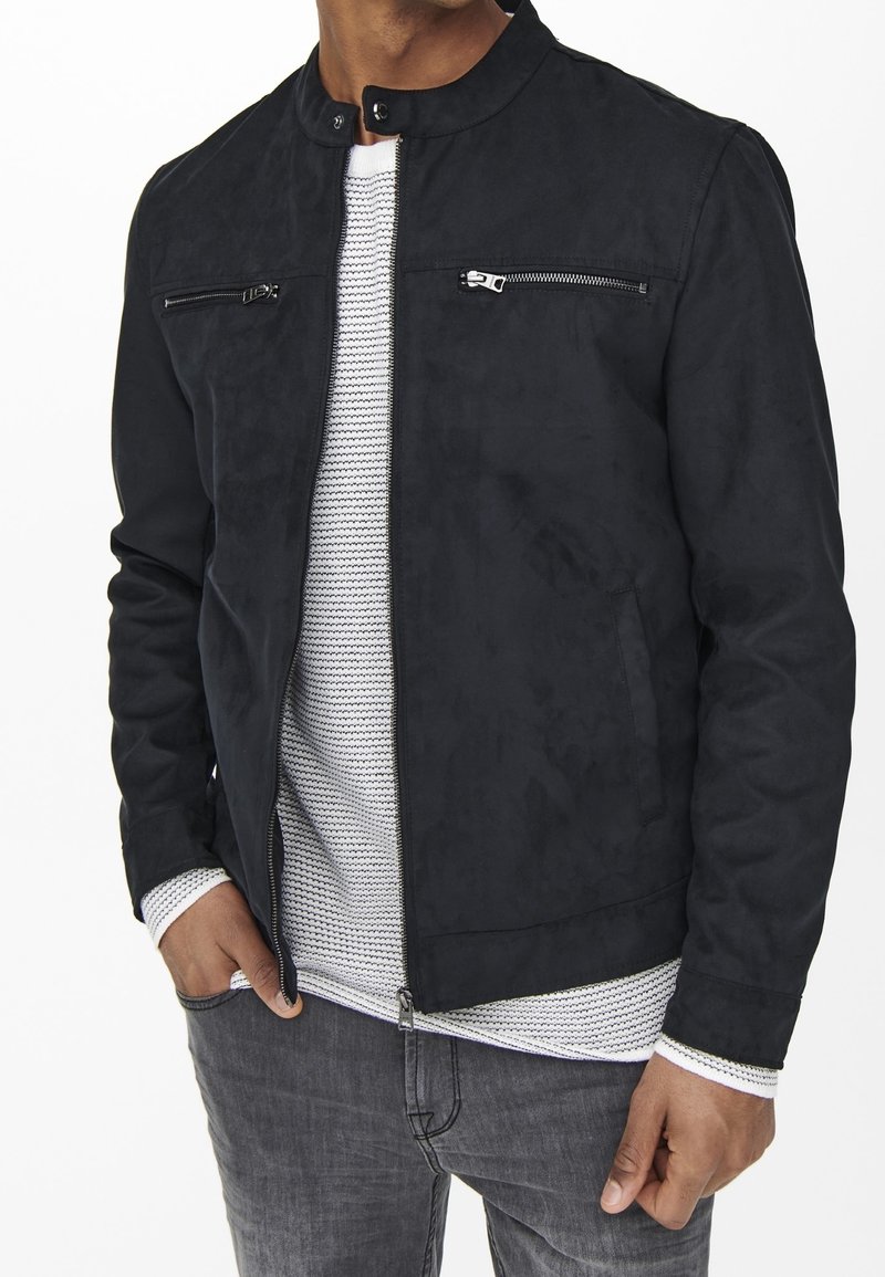 Giacca nera in suede con colletto alto, due tasche sul petto con zip e una trama liscia, abbinata a una camicia bianca a trama sottostante.