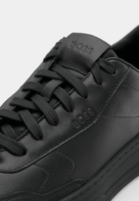 Zapatilla deportiva de cuero negro con cordones de tejido texturizado, que presenta un logo "BOSS" elevado en la lengüeta y costuras distintivas a lo largo de los lados.