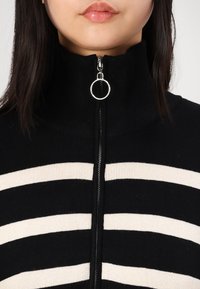 Suéter negro acanalado con cuello alto, con rayas horizontales en crema y un tirador de cremallera circular plateado en el cuello.