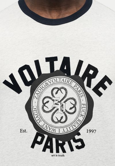 Μπλούζα με μαύρο κείμενο "Voltaire Paris", κυκλικό έμβλημα με πλεγμένα φίδια και μικρότερο κείμενο που περιλαμβάνει "Zadig & Voltaire Paris" και "Est. 1997."