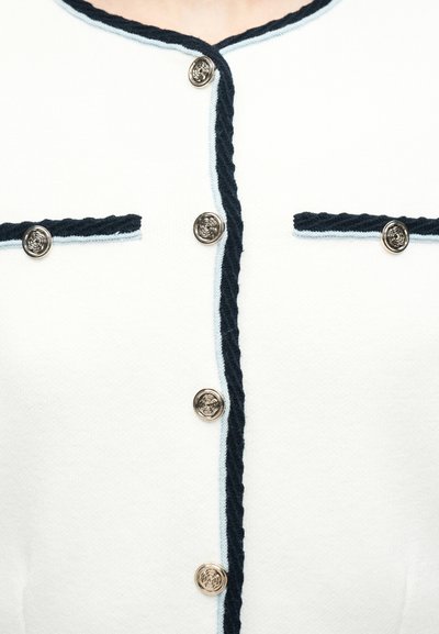 Λευκό ζακέτα με μαύρο περίγραμμα, τέσσερα διακοσμητικά ασημένια κουμπιά στο μπροστινό μέρος και δύο ταιριαστά κουμπιά στις τσέπες στο στήθος.