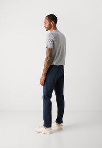 Mann mit Tattoos, der ein graues T-Shirt, marineblaue Hose und weiße Sneakers trägt und seitlich vor einem schlichten weißen Hintergrund steht.