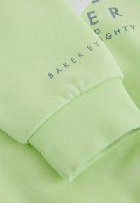 Lys grøn sweatshirt-stof med en tekstureret finish. Har en manchet og trykt tekst "BAKER BY MIGHTY" i gråt.