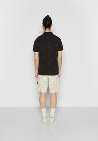 Person med hår i knut som bär en svart Calvin Klein Jeans skjorta, beige shorts, vita strumpor och vita sneakers, vänd mot en vit vägg.