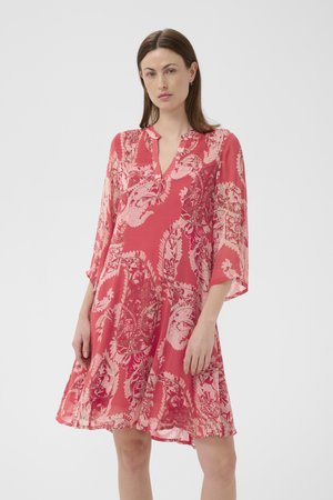 Suorahiuksinen ruskeatukkainen nainen käyttää löysää polvipituista korallinväristä mekkoa, jossa on vaaleanpunaisia ja punaisia paisley-kuvioita ja kolmen neljänneksen hihat, seisoo valkoista taustaa vasten.