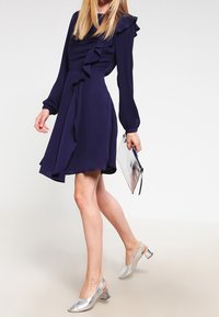 Mörkblå klänning med volangdetaljer, långa ärmar och asymmetrisk fåll. Matchad med silvriga slingback-klackar och en metallisk kuvertväska.