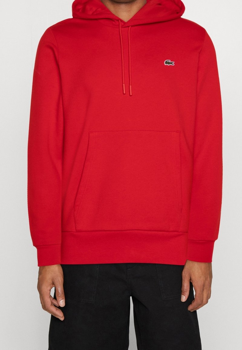 Homme portant un sweat à capuche rouge avec poche avant et cordons, associé à un pantalon noir, visible du cou jusqu'au milieu de la cuisse.