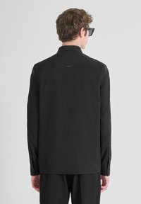Camisa negra de manga larga con corte recto, puños abotonados y cuello, hecha de una tela ligera. Branding minimal en la parte trasera.