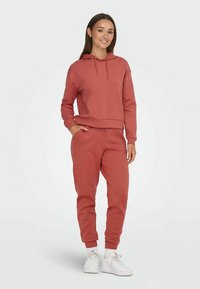 Różowa bluza z kapturem, ściągaczami na mankietach oraz pasujące joggery. Gładka faktura, zestawiona z białymi trampkami, na tle jasnego tła.