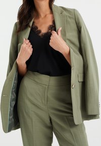 Olivgrön skräddarsydd kavaj i en texturerad tyg, parat med en svart topp med spetsdetaljer. Har knappar och slät foder.