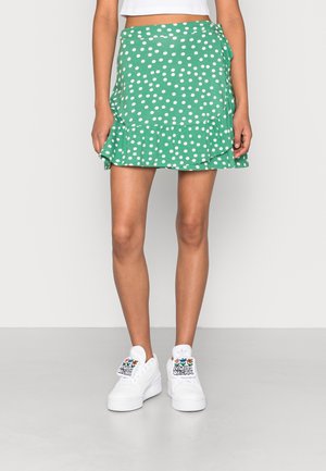 Femme portant une jupe portefeuille verte à pois blancs, un haut court blanc et des baskets blanches avec des patchs colorés "Always Original".
