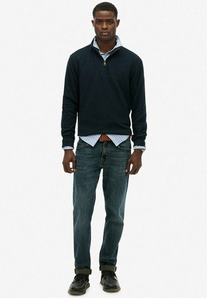 Sudadera negra con cremallera sobre una camisa de botones rayada en azul claro, combinada con jeans azules y zapatos negros. Un atuendo casual y en capas con colores contrastantes.