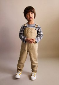Salopette en denim beige avec des bretelles ajustables, portée sur une chemise à manches longues gris et blanc à rayures, associée à des baskets blanches avec des accents jaunes.