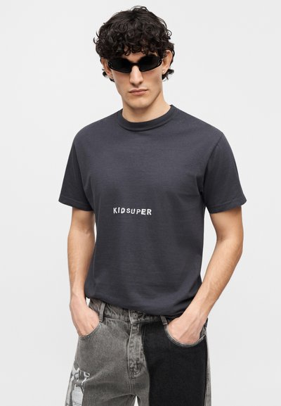 Σκούρο γκρι βαμβακερό T-shirt με στρογγυλή λαιμόκοψη και λευκό τυπωμένο λογότυπο "KIDSUPER". Συνδυασμένο με πολύχρωμο γκρι τζιν παντελόνι.