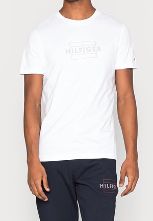 Mann trägt weißes T-Shirt und marineblaue Hose, beide mit bestickten "HILFIGER"-Logos in roten, umrandeten Rechtecken.