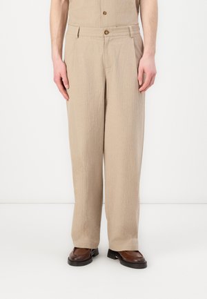 Pantaloni beige in tessuto testurizzato, con vestibilità rilassata, gambe dritte e chiusura con bottone in vita. Scarpe marroni completano il look.