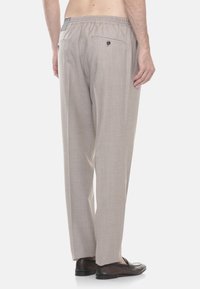 Pantaloni beige su misura con tasche posteriori a bottone indossati da una persona senza camicia, abbinati a mocassini in pelle marrone scuro.