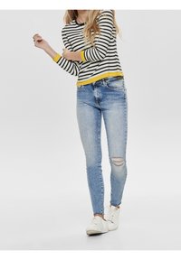 Randig långärmad tröja med gul nederkant, kombinerad med ljusblå skinny jeans med ett knäskär, och vita sneakers med texturerad detalj.