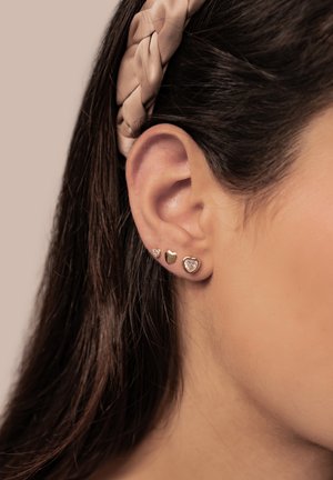 Gold-tone stud earrings in heart and round shapes, featuring clear stone accents. Worn on a pierced ear, with hair pulled back.