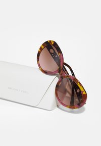 Ochelari de soare cu model țestoasă, cu o formă mare de tip cat-eye, lentile degradate de culoare maro și branding gravat. Însoțiți de un etui de protecție alb.
