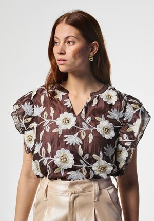 Frau mit langem roten Haar, die eine braune Bluse mit Blumenstickerei und beige hoch taillierte Hosen trägt, schaut zur Seite.