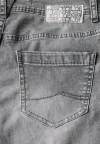 Grijze denim stof met een achterzak met subtiele stikdetails en een textuurleer etiket erboven, dat het merkontwerp toont.