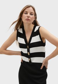 Czarna i biała, w paski, bez rękawów bluzka z dekoltem w kształcie V, dużymi złotymi guzikami i gładką fakturą. Projekt podkreśla kontrastujące kolory.