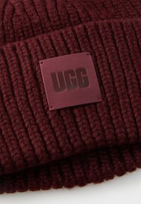 Bonnet tricoté bourgogne avec texture côtelée et un patch en cuir marron arborant le logo "UGG" embossé au centre.