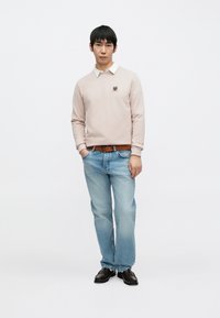 Lys rosa genser med rund hals og brodert logo, kombinert med lyseblå jeans og svarte sko, stående mot en hvit bakgrunn.