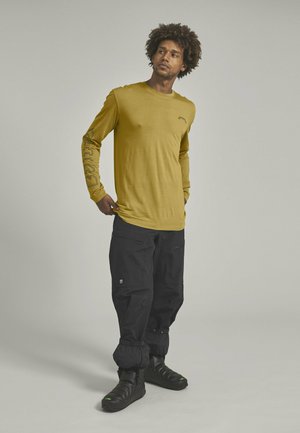 Langärmeliges senfgelbes Shirt mit grafischem Druck auf dem Ärmel, kombiniert mit schwarzen, lässigen Hosen und schwarzen Slippern.