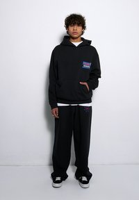 Czarna bluza z kapturem z przednią kieszenią oraz kolorowym logo na klatce piersiowej, w zestawie z pasującymi czarnymi spodniami dresowymi z graficznym wzorem.