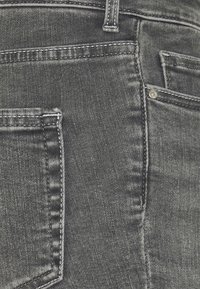 Tummanharmaat farkut, joissa on sileä rakenne, näkyvät ompeleet ja vakiopocket-muotoilu. Sisältää pienen metallirivettiyksityiskohdan.