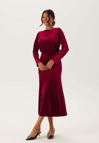 Långärmad klänning i djup burgundy sammet med figursydd överdel och flödande kjol, med rund halsringning och mjuk textur.