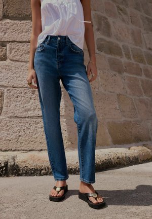 Kvinde iført blå jeans, hvid ærmeløs top og sorte sandaler med platåsål, stående foran en stenvæg.
